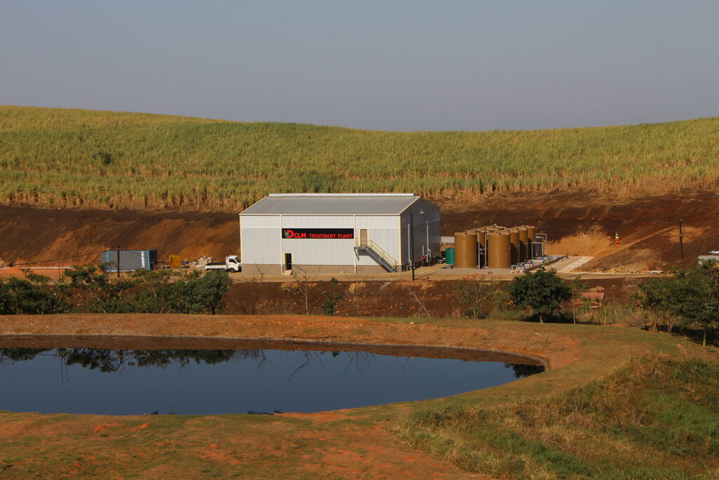 Steel-clad treatment plant at the hillside base with a long green field behind and a reflecting canal in the foreground.