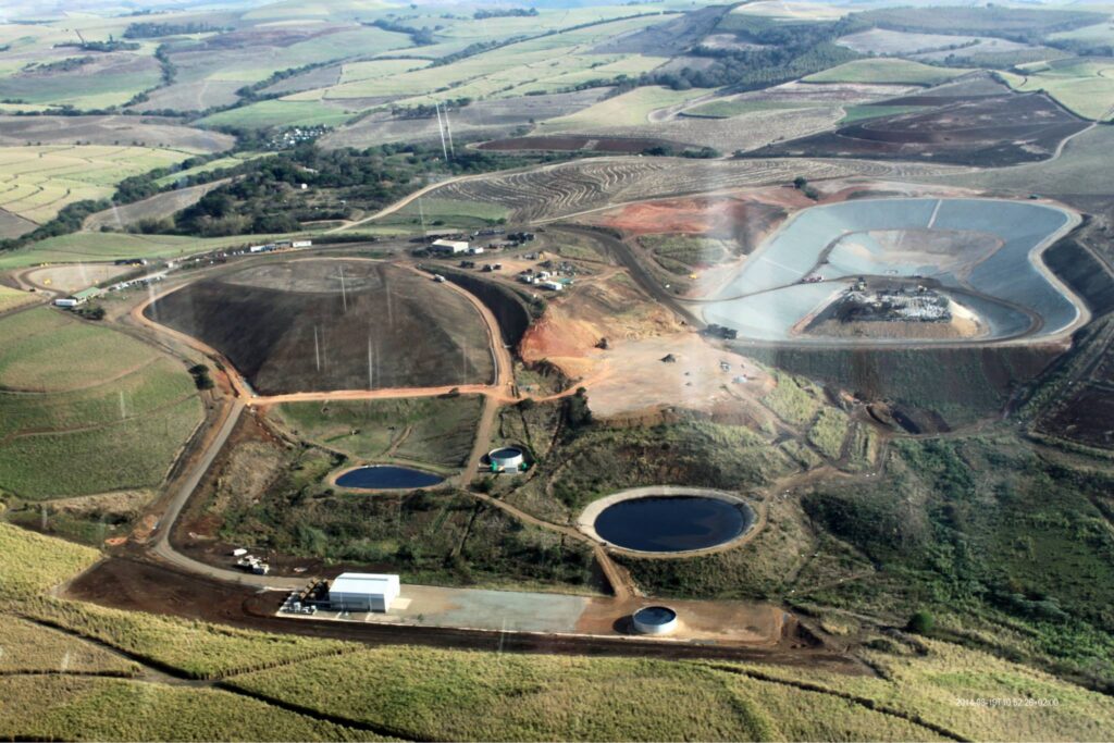 Aerial view of an open-pit mine with large circular excavations, roads, and scattered buildings amid surrounding farmland and rolling hills in the distance.