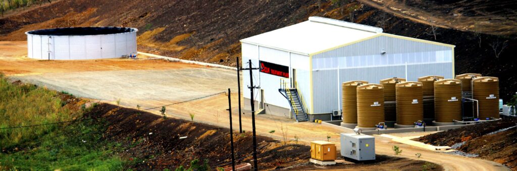 Industrial storage facility featuring a white metal building, a row of large brown cylindrical tanks, and a circular tank on a dirt yard amid a hilly landscape