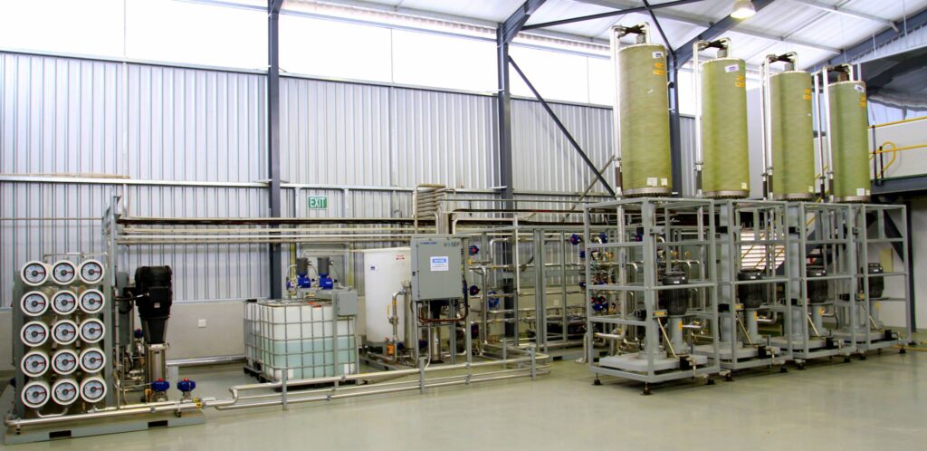 Industrial facility interior with vertical green processing tanks, extensive piping, and metal framework.
