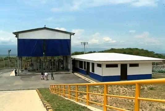 Outdoor industrial site with a blue canopy shelter over equipment, a white-and-blue building, and a yellow railing along a ramp in the foreground.