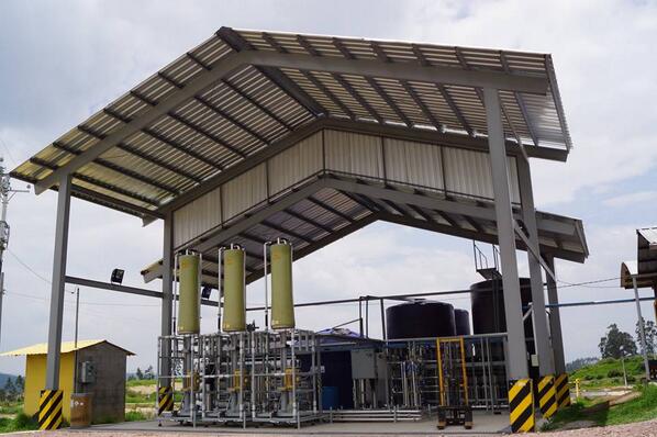Industrial processing station under a metal canopy with three tall yellow reactors and large storage tanks outside.