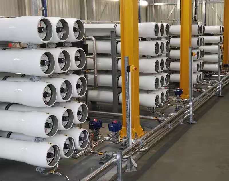 Row of white cylindrical membrane housings mounted on racks in a water treatment/filtration plant, with yellow support columns and stainless pipes nearby.