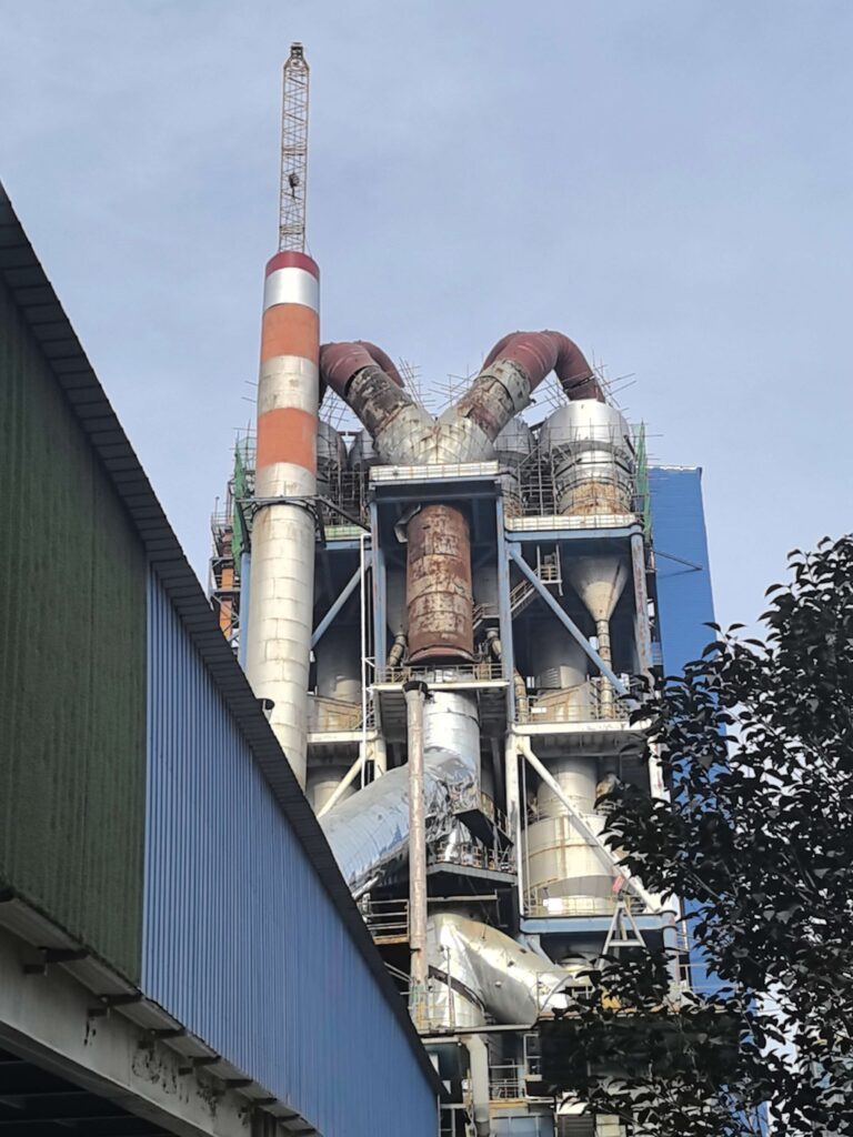 Industrial plant with a tall striped smokestack and network of pipes against a blue sky (left blue building visible).